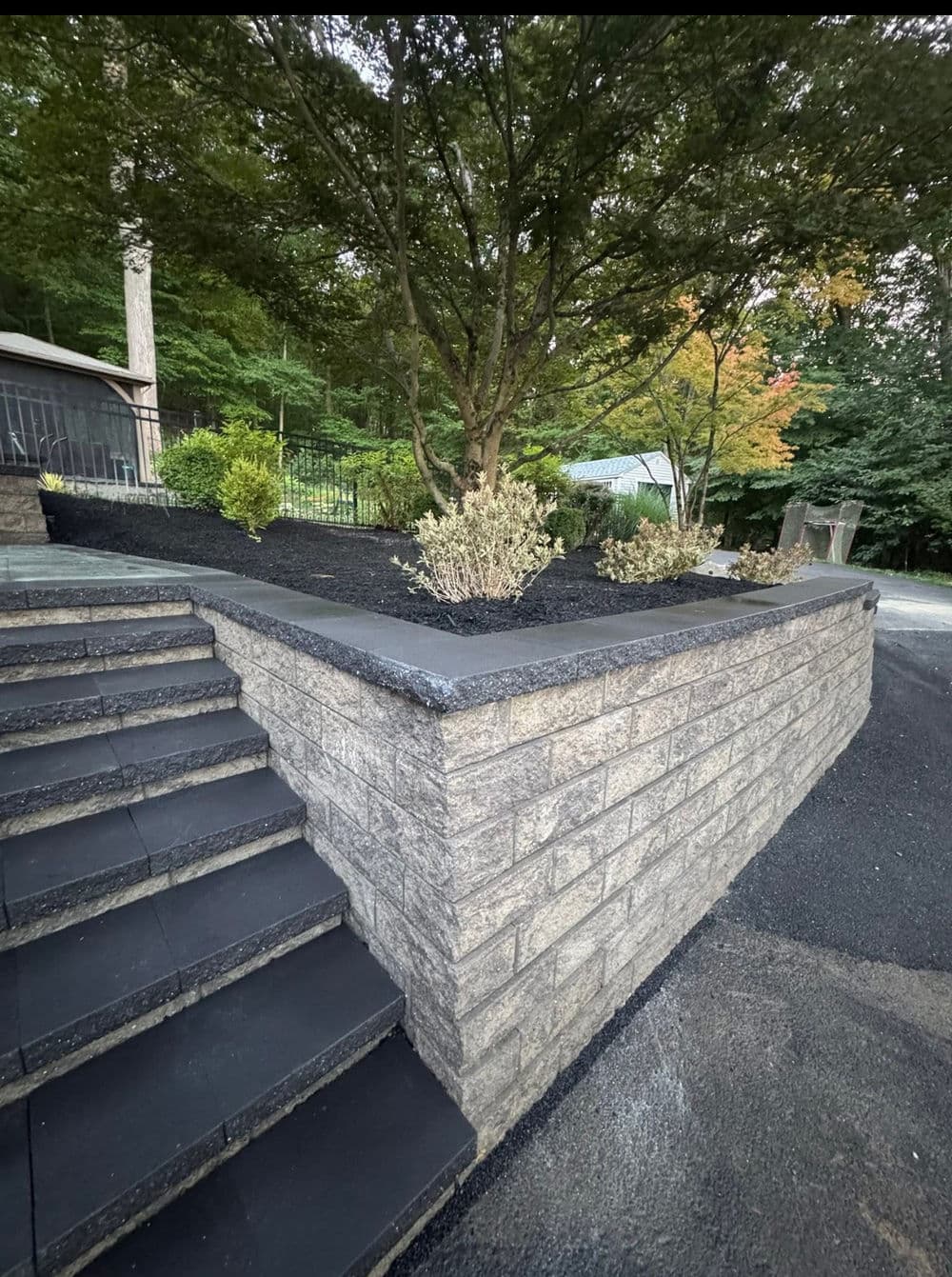 Stone retaining wall with steps and landscaped shrubs in a wooded outdoor setting.