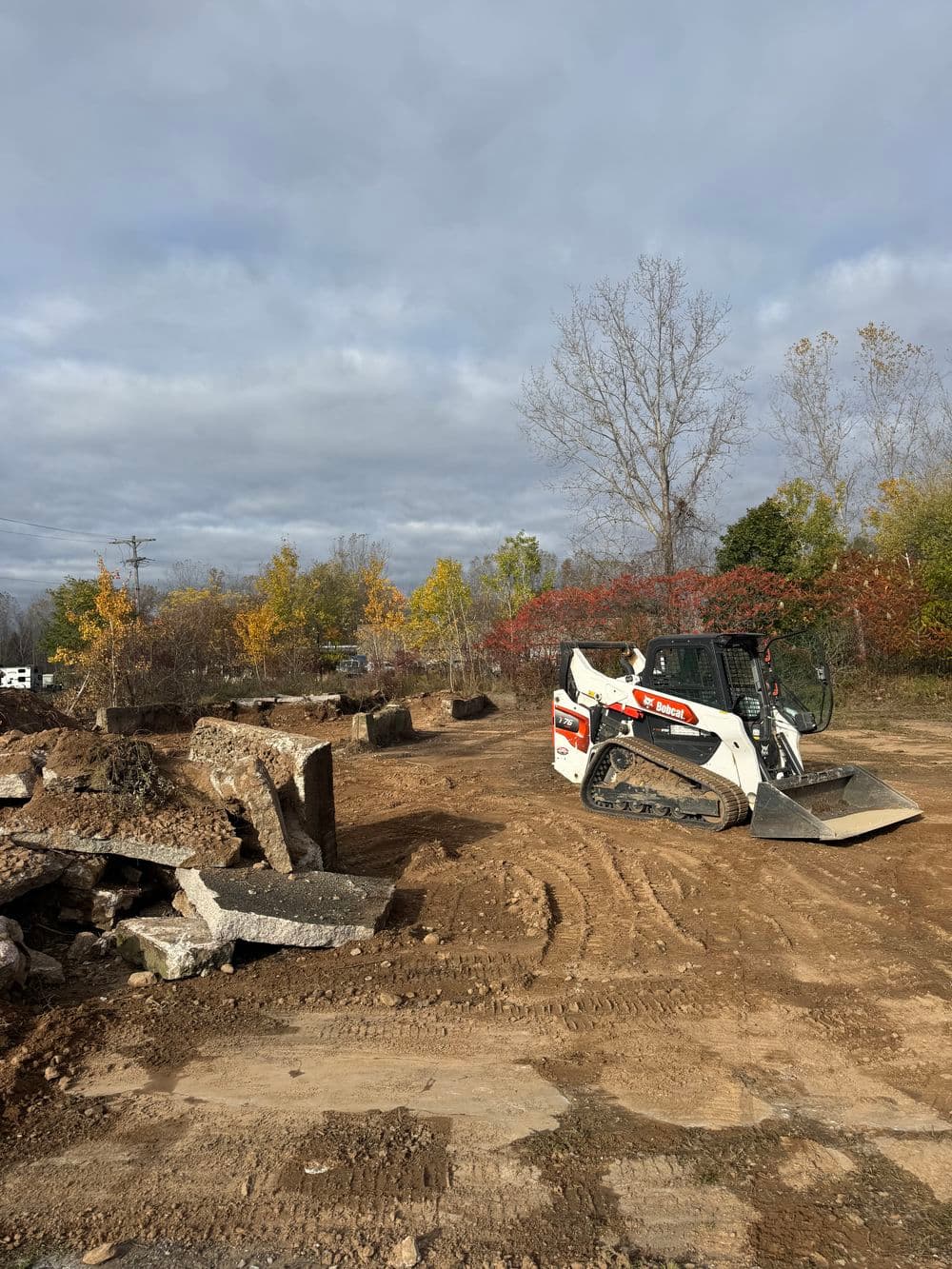 Bobcat excavator near rubble piles at a construction site with autumn trees in the background.
