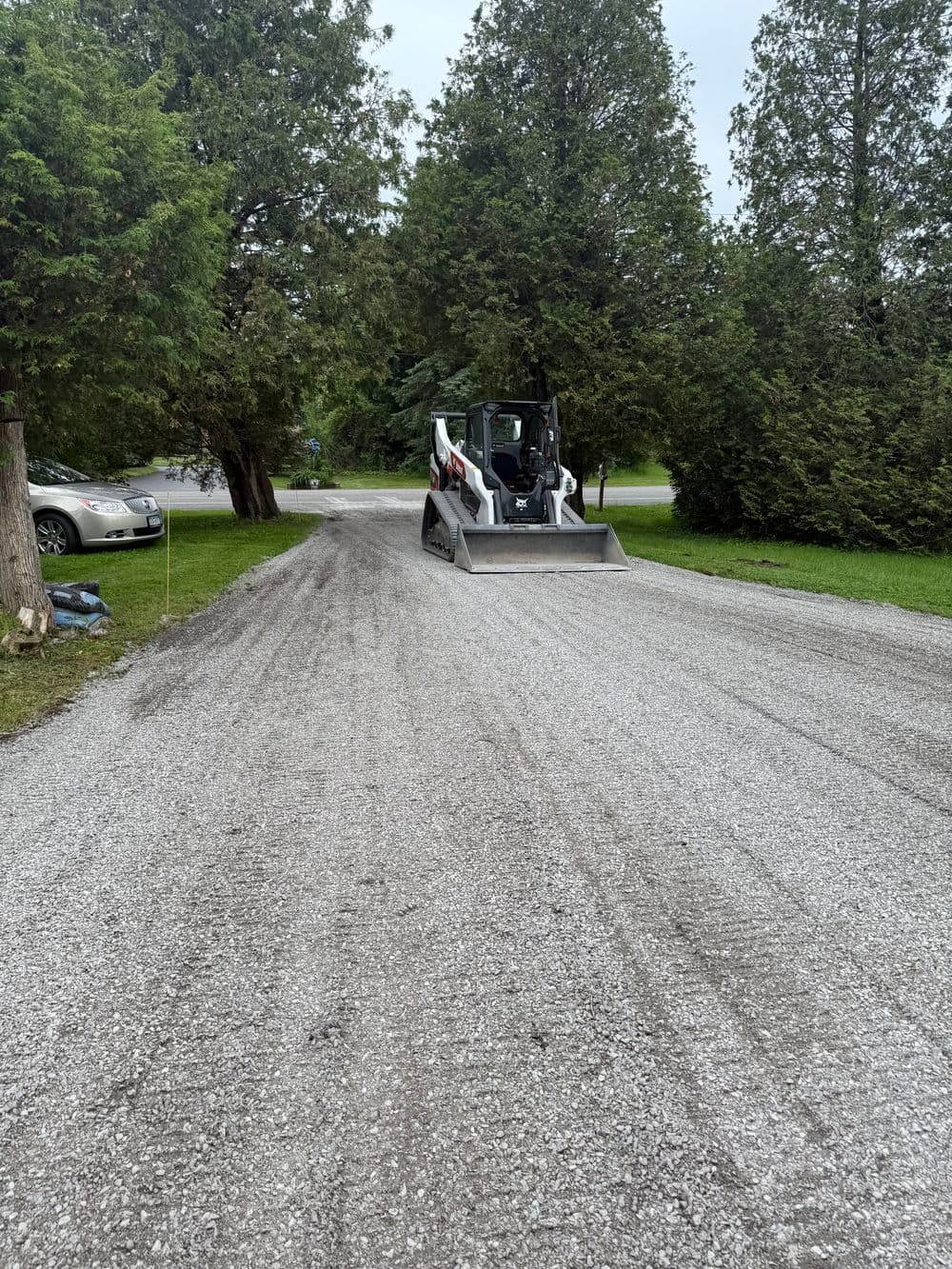 Bobcat machinery grading a gravel driveway surrounded by trees and parked cars.