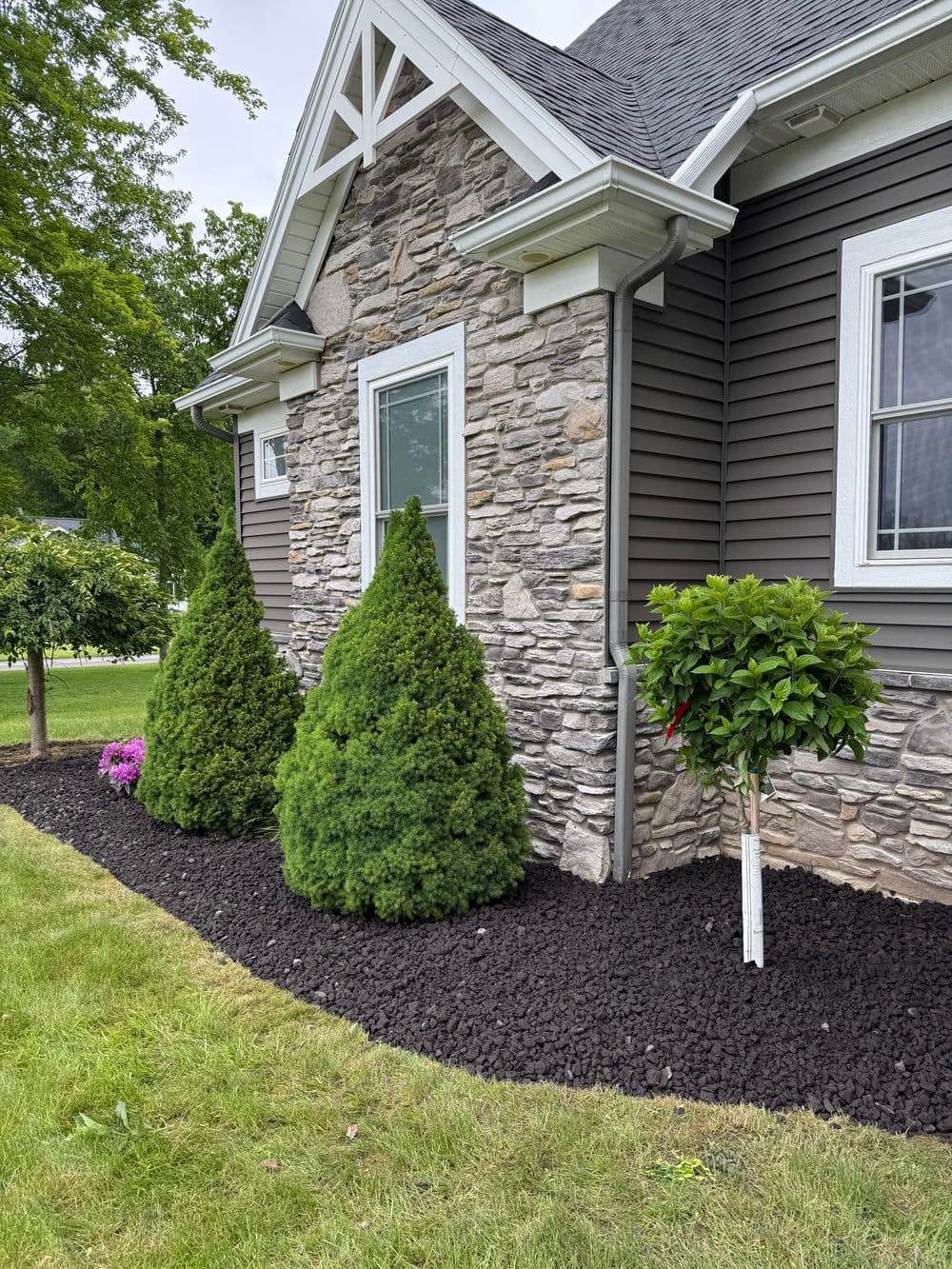 Well-maintained landscape featuring stone house, evergreen shrubs, and colorful flowers.