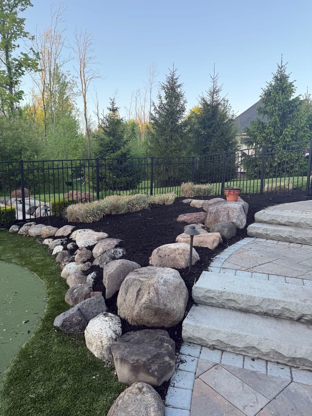 Garden landscape featuring rocks, mulch, and trees near a stone pathway and green turf.