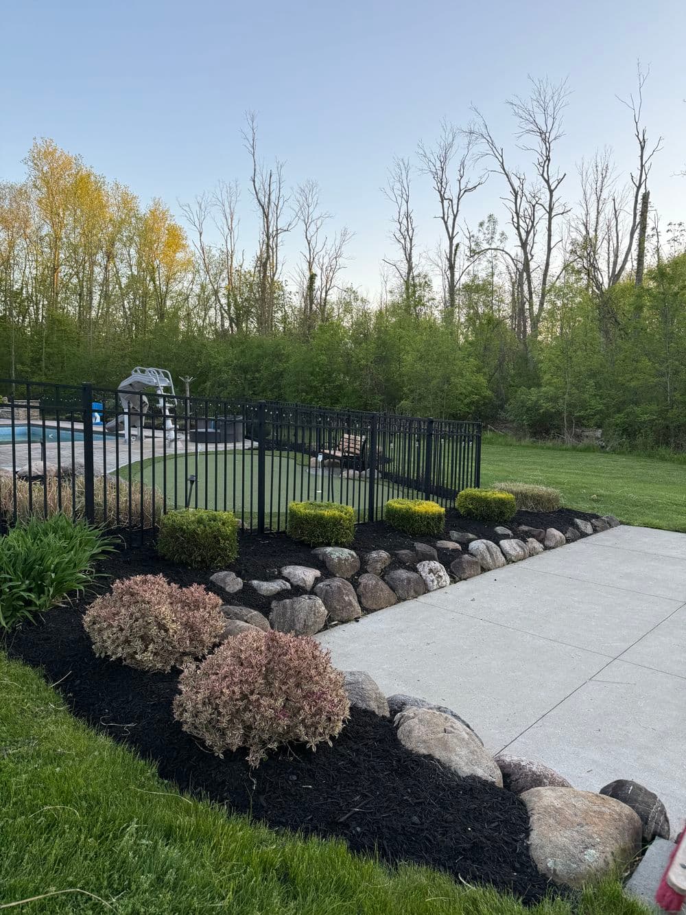 Lush green backyard with landscaped stone border, black fence, and a swimming pool area.